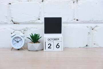 Wooden calendar with date October 26 and alarm clock with plant on table against brick wall background. Deadline, planning, business concept