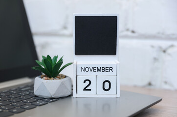 Wooden calendar with date November 20 and laptop, plant on table against brick wall background. Deadline, planning, business concept