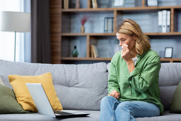 An older beautiful woman sits at home on the sofa with a laptop, cries, wipes her tears with a napkin. Financial problems, received bad news, a letter, watching a sad movie.