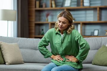 An elderly woman is sitting on the sofa at home, holding her stomach with her hands. Feels the pain...