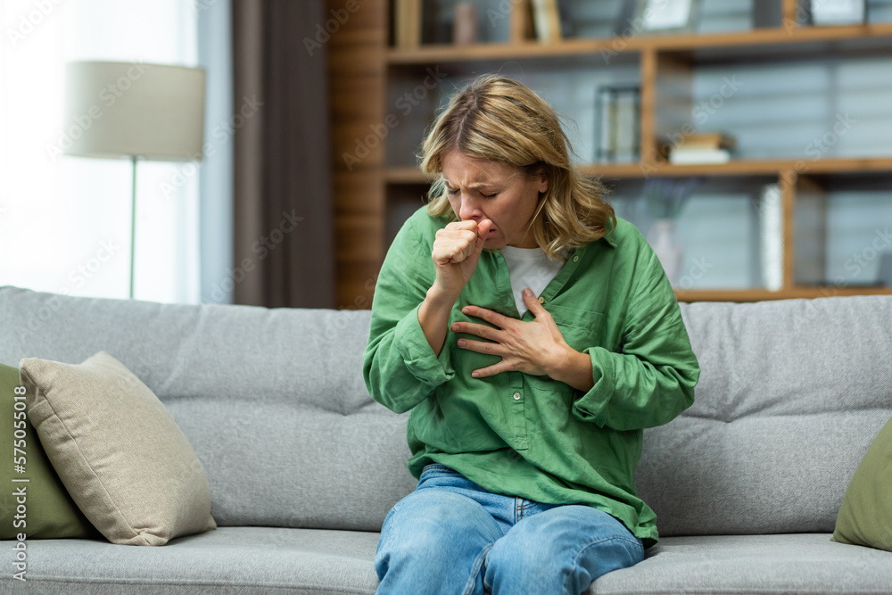 Wall mural Senior beautiful woman sitting sick on sofa at home. He coughs, covers his mouth with his hand, holds his chest. Feels pain, suffers from asthma, allergies, flu, cold.