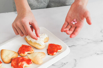 Hands of a caucasian teenage girl laying coffee cups heart sprinkles on not perfect freshly baked heart cookies .