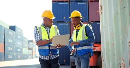 Two professional african engineers wearing safety uniform and hard hats working on laptop and holding a communication radio at cargo