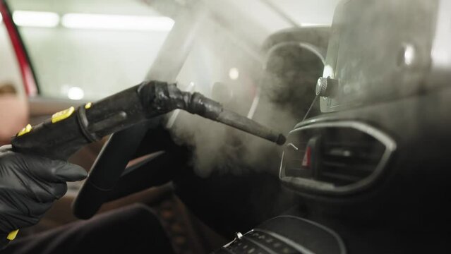 Close up of hands of man in black protective rubber gloves cleaning interior of the car with hot steam cleaner. Selective focus on guy hands. Auto cleaning service and detailing concept.