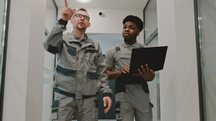 Two diverse installers in uniform walk hallway and discuss security cameras installation in modern...