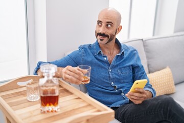 Young hispanic man using smartphone drinking whisky at home