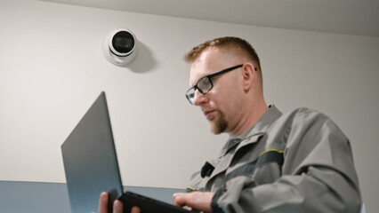Installer in uniform sets up security camera in office room using laptop. Adult Man checks CCTV...