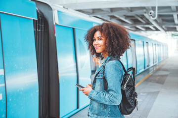 Happy young tourist African American woman with backpack and holding smartphone while at the train...