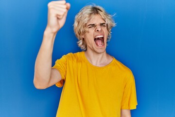 Young modern man standing over blue background angry and mad raising fist frustrated and furious while shouting with anger. rage and aggressive concept.