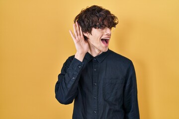 Fototapeta premium Young man wearing glasses over yellow background smiling with hand over ear listening an hearing to rumor or gossip. deafness concept.
