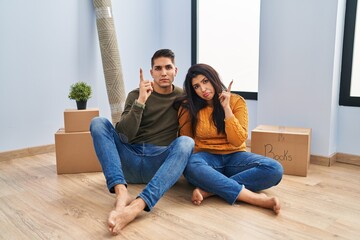 Young couple sitting on the floor at new home pointing up looking sad and upset, indicating...