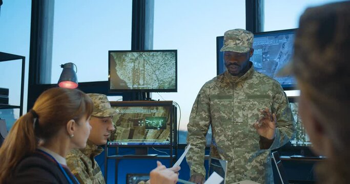 African American male head of militarian headquarters talking with report to Caucasian army staff while people studying and reading documents. Military training in office concept.