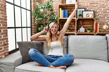 Young blonde woman listening to music and dancing sitting on sofa at home