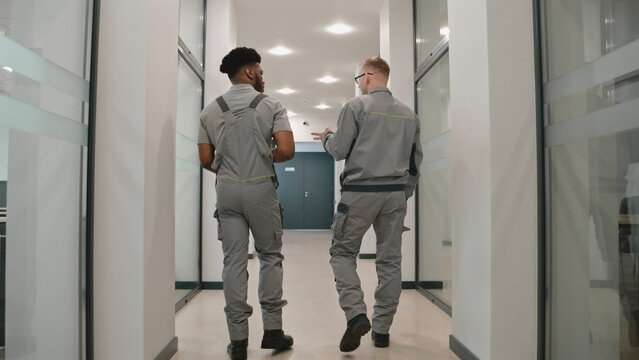 Back view of two diverse men in uniform walk hallway and discuss CCTV cameras or Internet installation in coworking office. Installers set up cameras using laptop. Concept of surveillance system.