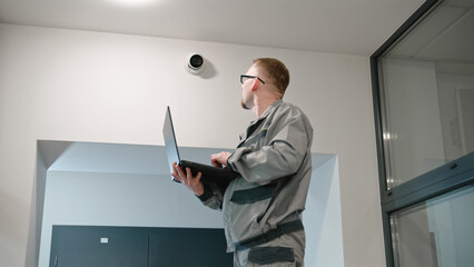 Installer in uniform sets up security camera in office room using laptop. Man in glasses checks...