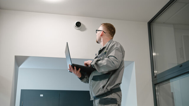 Installer In Uniform Sets Up Security Camera In Office Room Using Laptop. Man In Glasses Checks CCTV Cameras In Computer Program. Monitoring System. Concept Of Surveillance System And Privacy.