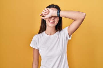 Young beautiful woman standing over yellow background covering eyes with arm smiling cheerful and funny. blind concept.