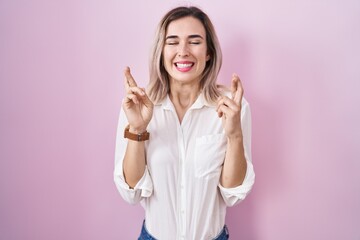 Young beautiful woman standing over pink background gesturing finger crossed smiling with hope and eyes closed. luck and superstitious concept.