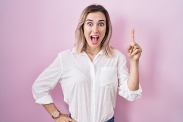 Young beautiful woman standing over pink background pointing finger up with successful idea. exited and happy. number one.