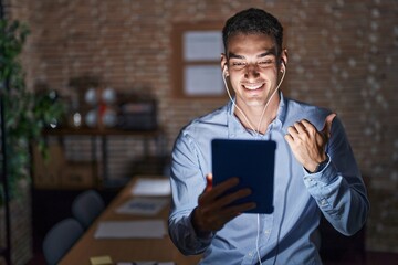 Handsome hispanic man working at the office at night pointing to the back behind with hand and thumbs up, smiling confident