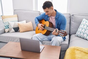 Young hispanic man having online classical guitar lesson sitting on sofa at home