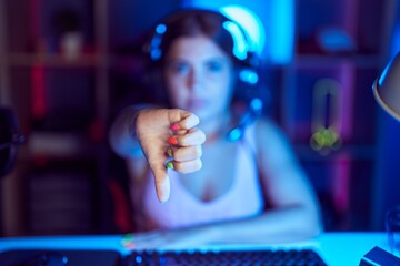 Young blonde woman playing video games wearing headphones looking unhappy and angry showing rejection and negative with thumbs down gesture. bad expression.