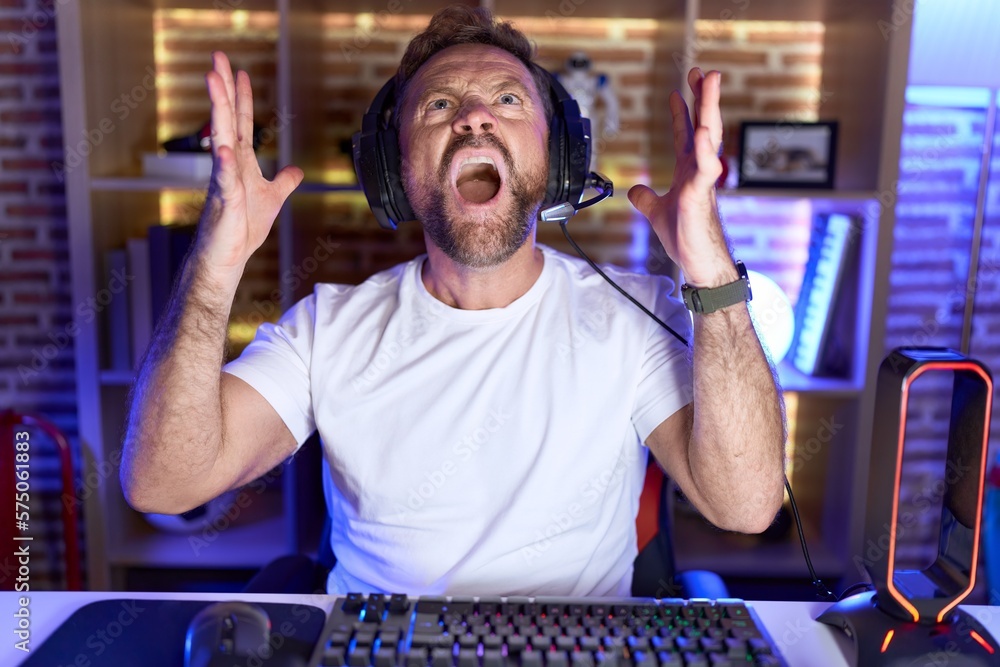 Sticker Middle age man with beard playing video games wearing headphones crazy and mad shouting and yelling with aggressive expression and arms raised. frustration concept.