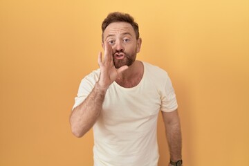 Middle age man with beard standing over yellow background hand on mouth telling secret rumor, whispering malicious talk conversation