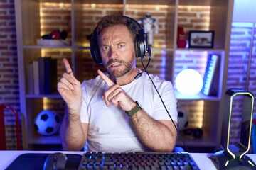 Middle age man with beard playing video games wearing headphones pointing aside worried and nervous...