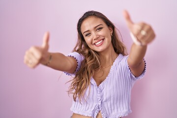 Young hispanic woman standing over pink background approving doing positive gesture with hand, thumbs up smiling and happy for success. winner gesture.