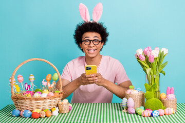 Photo of young excited surprised man use phone sit desk eggs easter holiday bunch fresh tulips delivery isolated on aquamarine color background