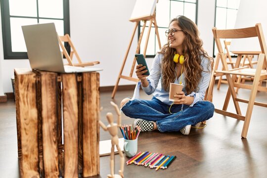 Young beautiful hispanic woman artist using smartphone drinking coffee at art studio