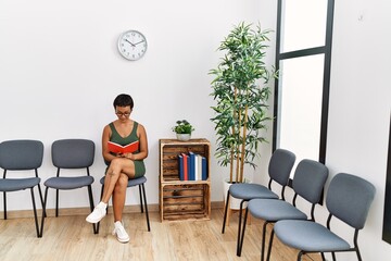 Young hispanic woman reading book sitting on chair at waiting room