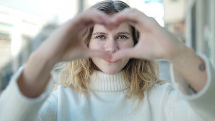 Young blonde woman doing heart gesture with hands over eyes at street