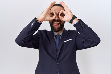 Hispanic man with beard wearing suit and tie doing ok gesture like binoculars sticking tongue out, eyes looking through fingers. crazy expression.