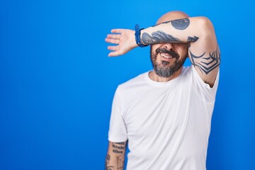 Hispanic man with tattoos standing over blue background covering eyes with arm, looking serious and...