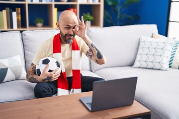 Hispanic man with tattoos watching football match hooligan holding ball on the laptop smiling happy doing ok sign with hand on eye looking through fingers
