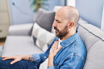 Young bald man sitting on sofa suffering heart attack at home