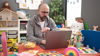 Young bald man preschool teacher using laptop looking watch at kindergarten