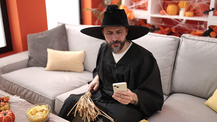 Young bald man wearing wizard costume using smartphone holding broom at home