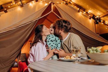Fototapeta na wymiar Happy family with lovely baby have dinner and spend time together in glamping on summer evening near cozy bonfire. Luxury camping tent for outdoor recreation and recreation. Lifestyle concept