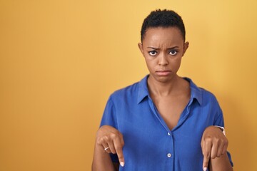 African american woman standing over yellow background pointing down looking sad and upset,...