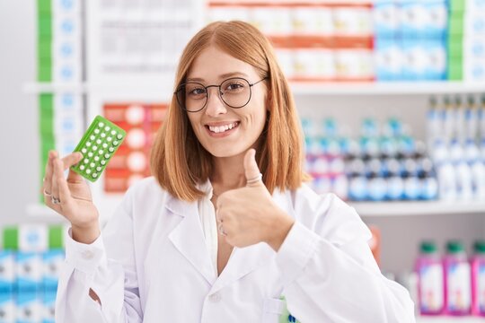 Young redhead woman working at pharmacy drugstore holding birth control pills smiling happy and positive, thumb up doing excellent and approval sign