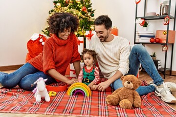 Couple and daughter playing with toys sitting by christmas tree at home