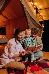 Obraz na płótnie Canvas Happy family with lovely baby playing and read book together in glamping on summer evening near cozy bonfire. Luxury camping tent for outdoor recreation and recreation. Lifestyle concept
