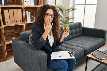 Young hispanic woman playing colorful puzzle cube intelligence game covering mouth with hand,...