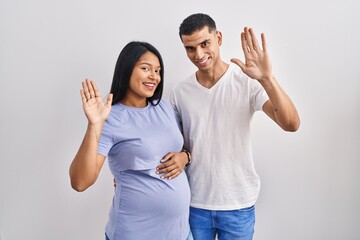 Young hispanic couple expecting a baby standing over background waiving saying hello happy and...