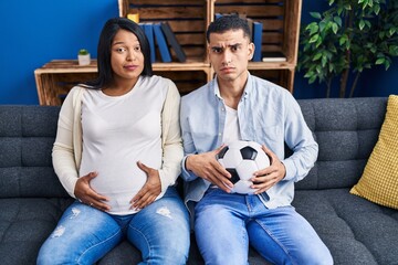 Young hispanic couple expecting a baby sitting on the sofa holding ball and tummy skeptic and...