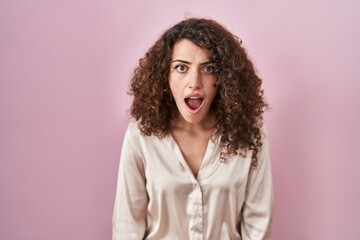 Hispanic woman with curly hair standing over pink background afraid and shocked with surprise and amazed expression, fear and excited face.