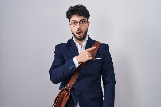 Hispanic man with beard wearing business clothes surprised pointing with finger to the side, open mouth amazed expression.